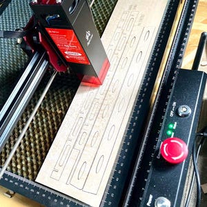 May include: A laser cutter in operation, focused on a light-colored wooden plank. The machine's red laser head is actively cutting out a design with oval and rectangular shapes. The machine has a black and red casing with safety labels.