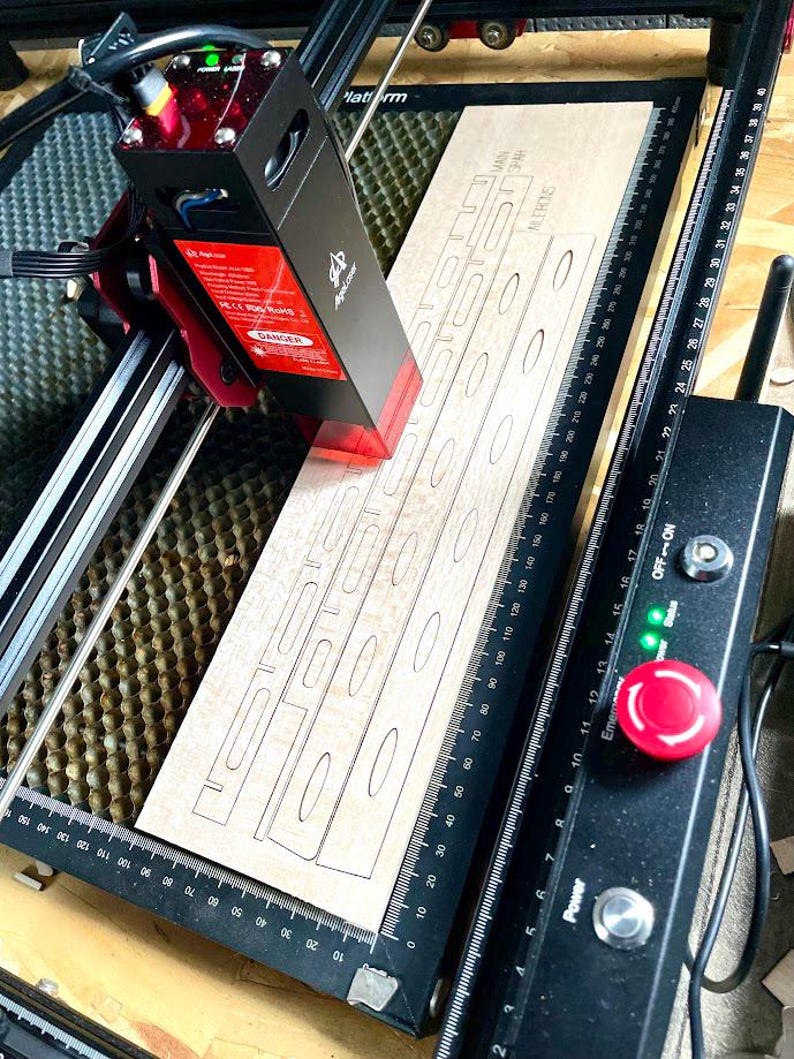 May include: A laser cutter in operation, focused on a light-colored wooden plank. The machine's red laser head is actively cutting out a design with oval and rectangular shapes. The machine has a black and red casing with safety labels.