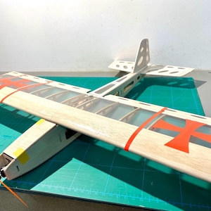 May include: A wooden model airplane with a clear wing design and orange propeller. The wings feature red crosses and red stripes. The model is set on a green surface.
