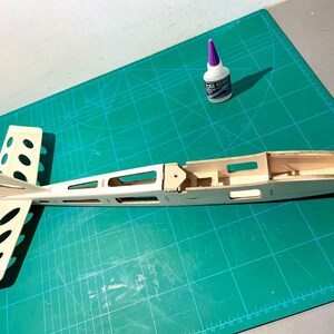 May include: A partially constructed wooden model airplane, featuring a fuselage and tail section with cut-out details. A bottle of glue with a purple cap sits nearby on a green cutting mat, suggesting a hobby or craft project.