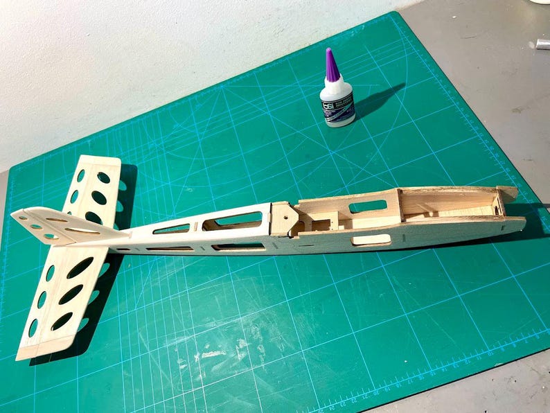 May include: A partially constructed wooden model airplane, featuring a fuselage and tail section with cut-out details. A bottle of glue with a purple cap sits nearby on a green cutting mat, suggesting a hobby or craft project.