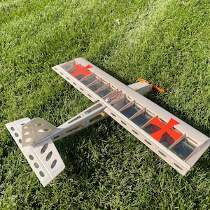 May include: A model airplane with a silver frame, red cross markings, and an orange propeller rests on green grass. The wings have a transparent covering, and the tail features multiple oval cutouts.