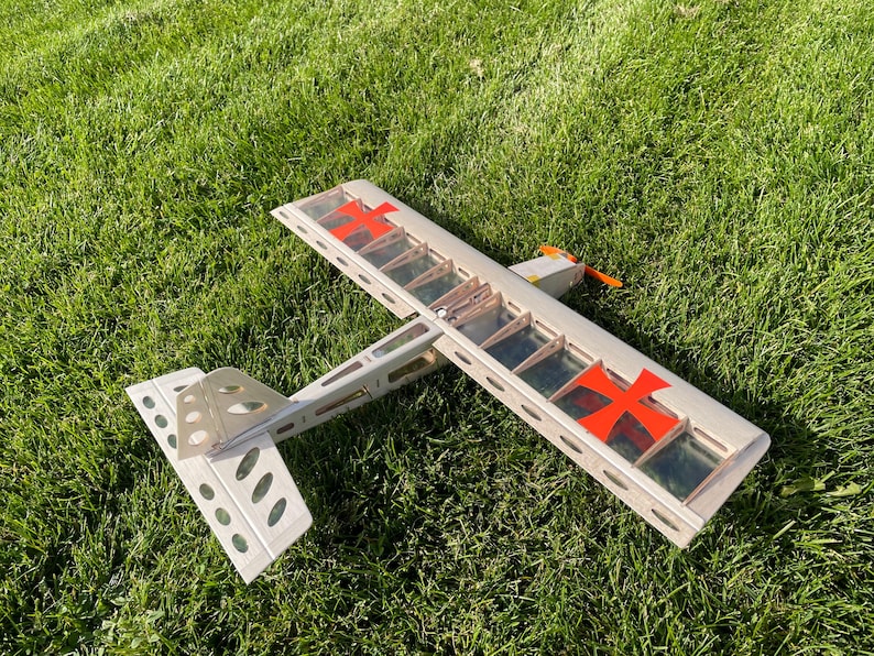May include: A model airplane with a silver frame, red cross markings, and an orange propeller rests on green grass. The wings have a transparent covering, and the tail features multiple oval cutouts.