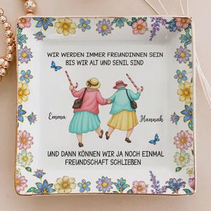 May include: Square ceramic trinket dish with a floral border. The center features an illustration of two elderly women holding candy canes. Text in German reads "We will always be friends until we are old and senile."