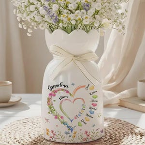 May include: White ceramic vase with a scalloped top, tied with a cream ribbon. The vase features a floral heart design with the words "Grandma" and "Mom", along with names. The vase is filled with white flowers.
