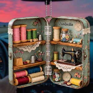 May include: A vintage-style sewing kit ornament in a teal metal case, open to reveal spools of thread in various colors, buttons, pins, a thimble, and a miniature sewing machine. The name "Isabella" is written on the case.