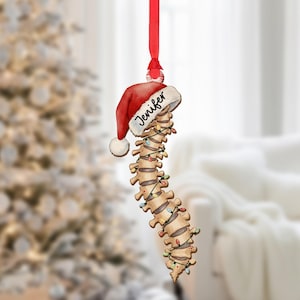 May include: A Christmas ornament shaped like a spine, adorned with a Santa hat and string lights. The hat is red with a white trim and the name "Jenifer" is written on it. The spine is tan and the lights are multicolored.