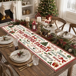 May include: A long rectangular white fabric table runner featuring the red text THE SMITHS FAMILY in the center, surrounded by various names in black script, colorful illustrations of Christmas trees, gifts, bows, and holly berries.
