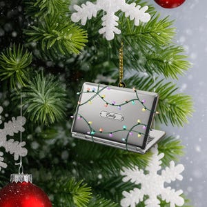 May include: Silver laptop Christmas ornament with colorful string lights. The ornament has the name "Emily" on it. The ornament is hanging on a Christmas tree with red ornaments and white snowflakes.
