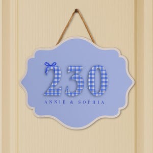 May include: A light blue, scalloped-edge sign with the number "230" in a blue and white plaid pattern. A blue bow is above the number. The names "ANNIE & SOPHIA" are printed below the number. The sign hangs from a brown string.