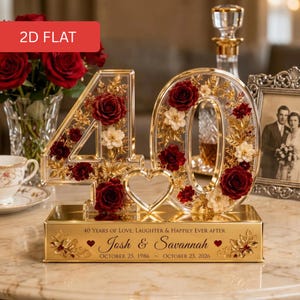 Peut inclure: Un présentoir en acrylique transparent en forme de chiffre 40 rempli de roses rouge foncé, de fleurs blanches et de feuilles dorées, avec un cœur central. La base dorée porte l'inscription 40 YEARS OF LOVE, LAUGHTER & HAPPILY EVER AFTER, Josh & Savannah et les dates.