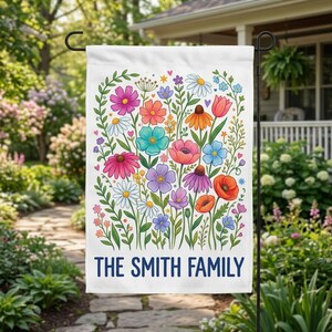 May include: A white rectangular garden flag featuring a colorful illustration of various wildflowers in shades of pink, orange, blue, purple, and yellow. The text THE SMITH FAMILY is printed in bold blue capital letters at the bottom.