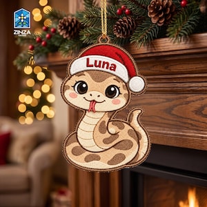Könnte beinhalten: Ein hängender Weihnachtsbaumschmuck in Form einer Cartoon-Schlange mit hellbraunen und dunkelbraunen Flecken, die eine rot-weiße Weihnachtsmütze mit dem in roter Schrift gestickten Namen Luna trägt. Der Anhänger hat eine goldene Schlaufe.