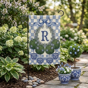 May include: A garden flag with a blue and white chequered background. The flag features a wreath with the letter "R" in the centre, and topiary trees in blue pots. A decorative bow is at the top of the flag.