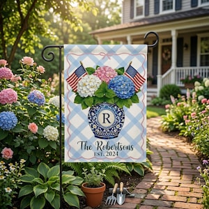 May include: A garden flag with a blue and white checkered pattern, featuring a vase with pink, white, and blue hydrangeas and two American flags. The flag reads "The Robertsons EST. 2024". The flag is displayed in a garden.