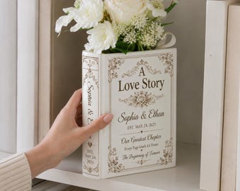 May include: A white ceramic book-shaped vase featuring gold floral filigree and the text A Love Story, Sophia & Ethan, EST. MAY 24, 2025, Our Greatest Chapter, Every Page Leads Us Home, The Beginning of Forever, filled with white roses and greenery.