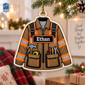 Può includere: Un ornamento piatto da appendere a forma di giacca da lavoro marrone con strisce riflettenti arancioni e grigie. La giacca presenta un'etichetta nera con il testo bianco Ethan e tasche contenenti un martello, un metro e due cacciaviti.