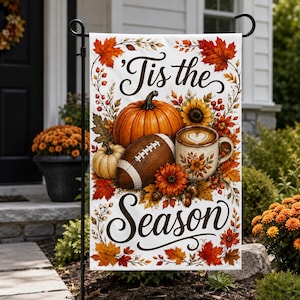 以下が含まれることがあります： 黒い筆記体で「Tis the Season」と書かれた白いガーデンフラッグ。大きなオレンジ色のカボチャ、小さな白いカボチャ、アメリカンフットボール、ラテアートが施された陶器のマグカップ、秋の葉、ヒマワリが描かれています。