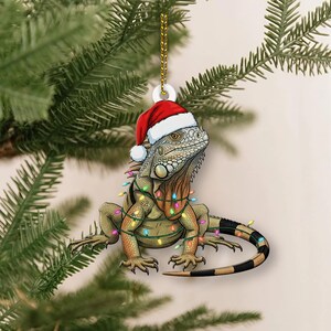 Könnte beinhalten: Weihnachtsschmuck mit einer detaillierten Illustration einer Leguan, der einen Weihnachtsmannhut trägt und mit bunten Lichterketten geschmückt ist. Der Leguan hat einen grün-braunen Körper und einen schwarz-weiß gestreiften Schwanz. Der Schmuck hängt an einer goldenen Kette.