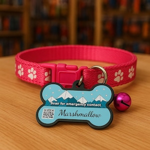 Peut inclure: Un collier rose vif pour chien avec des empreintes de pattes blanches et une boucle rose. Une étiquette en forme d'os avec un motif de montagne et le nom "Marshmallow" est attachée, ainsi qu'une petite cloche rose. L'étiquette comprend un code QR et le texte "Scan for emergency contact."
