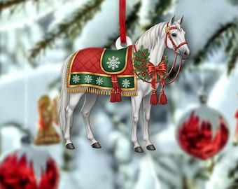 May include: A white horse Christmas ornament with a red bridle and a festive blanket. The blanket is red with a green border and snowflake accents. A red ribbon is attached for hanging.