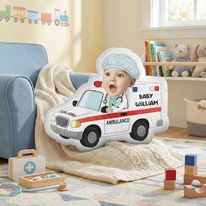 May include: A white and red ambulance-shaped pillow with a baby's face and the name "Baby William." The baby is wearing a medical cap and stethoscope. The pillow is on a cream-colored blanket, with a toy doctor's kit and blocks nearby.