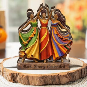 May include: A wooden sculpture of three women in colorful dresses, arms raised in celebration. The dresses are green, red, and rainbow-striped. The base reads "Black. Strong. Celebrated."