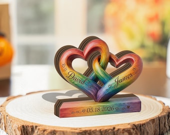 May include: A wooden rainbow-colored heart sculpture with two intertwined hearts, personalized with the names "David" and "James" and the date "05.18.2026". The sculpture sits on a wooden base and is displayed on a wooden slice.