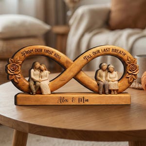 Könnte beinhalten: Eine Holzskulptur mit einem Unendlichkeitssymbol und dem Text "From our first kiss" und "Till our last breath." Die Skulptur zeigt zwei Figurenpaare, eines jung und eines älter, sowie die Namen "Alex & Mia." Auf beiden Seiten sind Rosen geschnitzt.