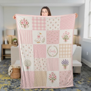 Pode incluir: Uma manta retangular macia com um design de patchwork em tons de rosa, branco e bege. O padrão inclui ilustrações de ramos, um pato com um laço rosa e uma coroa central contendo o nome Emily em letra cursiva cor-de-rosa.