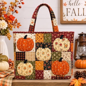 Op de afbeelding: Een gewatteerde draagtas met een patchworkontwerp van herfststoffen in tinten oranje, rood, groen en zwart. De tas is voorzien van vier stoffen pompoenapplicaties met bloemmotieven en twee stevige stoffen handvatten aan de bovenzijde.
