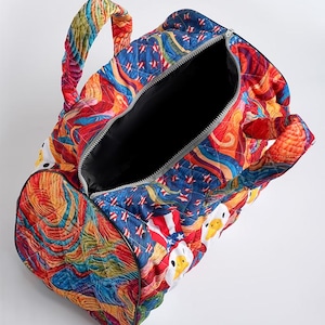 Puede incluir: Una bolsa de viaje acolchada con un patr&oacute;n multicolor vibrante de formas abstractas arremolinadas y cabezas de pato blancas con sombreros patri&oacute;ticos de estrellas. La bolsa tiene dos asas acolchadas y un forro interior de color negro.