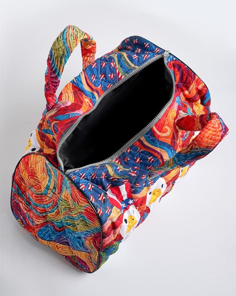 Puede incluir: Una bolsa de viaje acolchada con un patr&oacute;n multicolor vibrante de formas abstractas arremolinadas y cabezas de pato blancas con sombreros patri&oacute;ticos de estrellas. La bolsa tiene dos asas acolchadas y un forro interior de color negro.