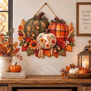 May include: A wall hanging decoration featuring three pumpkins in green, orange plaid, and white with floral patterns. The arrangement is accented with autumn leaves, wheat stalks, red berries, and acorns, all suspended by a rustic twine rope.