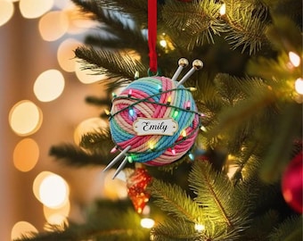May include: A Christmas tree ornament shaped like a ball of yarn, with knitting needles and a string of lights. The yarn is multi-colored, with blue, pink, and green hues. A small tag with the name "Emily" is attached. The ornament hangs from a red ribbon.