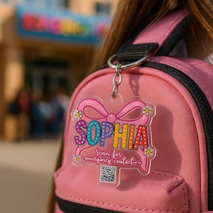 Könnte beinhalten: Ein rosa Rucksack mit einem transparenten, dekorativen Schlüsselanhänger. Der Schlüsselanhänger trägt den Namen "SOPHIA" in bunten Buchstaben, eine Schleife und den Text "Scan for emergency contact" mit einem QR-Code.