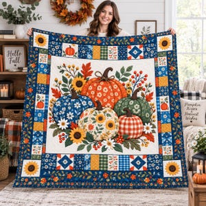 May include: A rectangular quilt featuring a central panel with four pumpkins in orange, blue, green, and white with floral and plaid patterns, surrounded by sunflowers and autumn leaves. The border consists of blue, yellow, and orange patchwork squares.