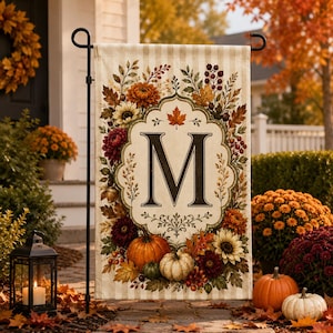 May include: A rectangular garden flag featuring a cream and beige striped background with a central ornate frame containing the letter M. The design is surrounded by a botanical border of autumn leaves, sunflowers, chrysanthemums, and pumpkins.