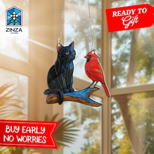 Puede incluir: Un atrapasol de vidrio coloreado con un gato negro y un cardenal rojo posados en una rama. El atrapasol está colgado de una cadena plateada y tiene una rama azul y marrón.