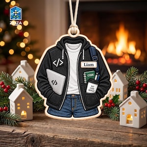 Può includere: Un ornamento da appendere a forma di felpa nera con cappuccio sopra una maglietta bianca e jeans blu. La felpa presenta un'icona di codice, un cartellino con il nome Liam, un taccuino verde con scritto CODE e un bicchiere con EAT CODE REPEAT.