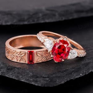 May include: A set of two rose gold engagement rings with a red ruby gemstone and white diamonds. The band of the larger ring features a floral design.