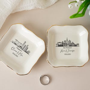 Puede incluir: Dos cuencos de cerámica blanquecina para anillos con bordes dorados, con ilustraciones de horizonte negro y texto personalizado. Uno dice "Singapur" y el otro "San Francisco". Un anillo plateado descansa entre los cuencos.