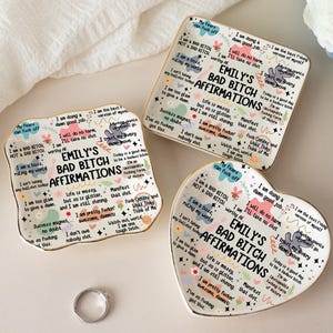 May include: Three decorative ceramic dishes with gold trim, featuring the text "EMILY'S BAD BITCH AFFIRMATIONS" and various affirmations. The dishes are in square, heart, and irregular shapes, with colorful floral and text designs.