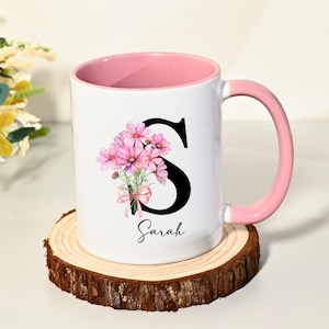 以下が含まれることがあります： ピンク色の内側とハンドルが付いた白いセラミックマグ。マグには、ピンクの花束と筆記体で「Sarah」という名前が付いた黒い文字「S」が描かれています。マグは木製のコースターの上に置かれています。
