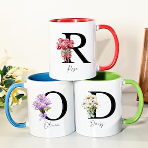 May include: Three white ceramic mugs stacked, each with a different initial and floral design. The mugs have coloured rims and handles: red, blue, and green. The mug with the letter "R" has a red rim and handle, with the name "Rose" below.