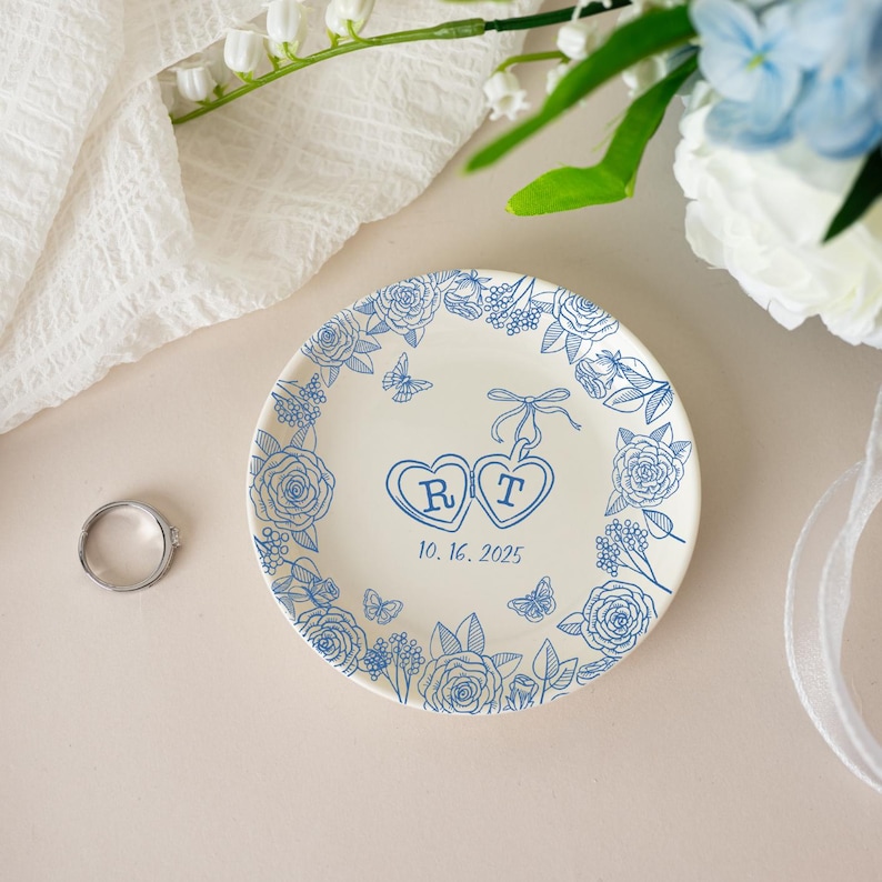 K&ouml;nnte beinhalten: Ein wei&szlig;es Keramik-Ringtablett mit blauem Blumendesign und zwei herzf&ouml;rmigen Initialen. Das Tablett ist mit Rosen und Schmetterlingen verziert, und das Datum 10.16.2025 ist unter den Herzen aufgedruckt. Ein silberner Ring liegt auf der Oberfl&auml;che.