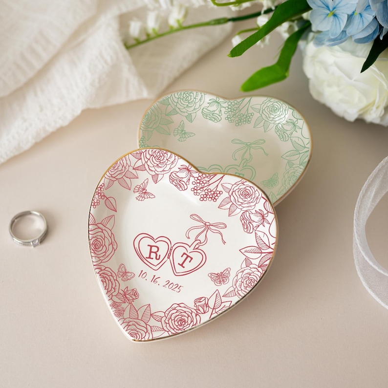 K&ouml;nnte beinhalten: Zwei herzf&ouml;rmige Ringschalen. Eine ist wei&szlig; mit einem roten Blumendesign, Initialen und einem Datum. Die andere ist wei&szlig; mit einem gr&uuml;nen Blumendesign. Beide haben goldene R&auml;nder. Ein silberner Ring liegt in der N&auml;he.