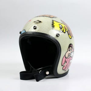 May include: Cream-colored motorcycle helmet with cartoon character graphics. The helmet has a black interior and trim, with a black chin strap. The helmet has the text "SBC" on the top.