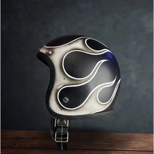 May include: Black motorcycle helmet featuring a flame design in white and grey. The helmet has a chin strap with a black buckle. The helmet is on a wooden surface.