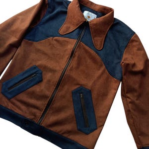Puede incluir: Chaqueta marrón y azul marino con cremallera. La chaqueta presenta un cuello ancho, dos bolsillos delanteros con cremalleras y detalles en azul marino en el cuello, los bolsillos y los puños. Fabricada en un material suave similar al ante.